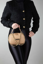 A person wearing a black outfit, holding a crème caramel AVAIA Petit, using the gold chain strap, twisted in a handheld style.