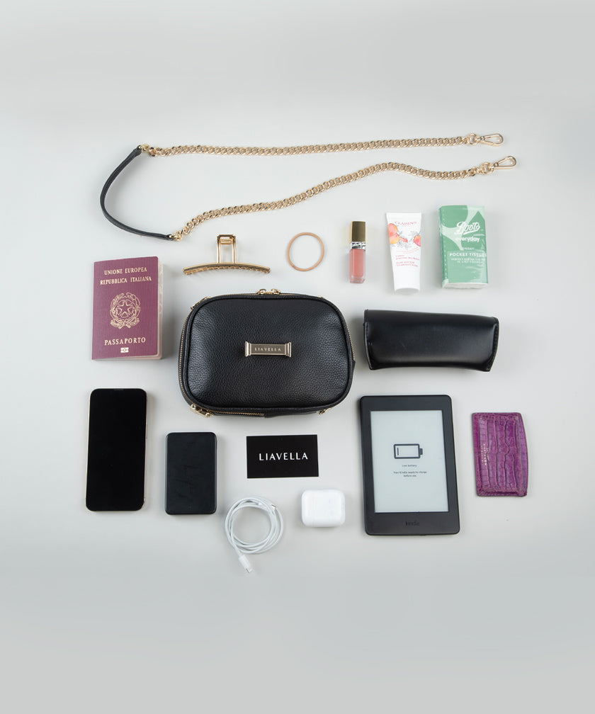Collection of travel essentials including a noirAVAIA Petit bag, phone, passport, and other items on a light gray background.
