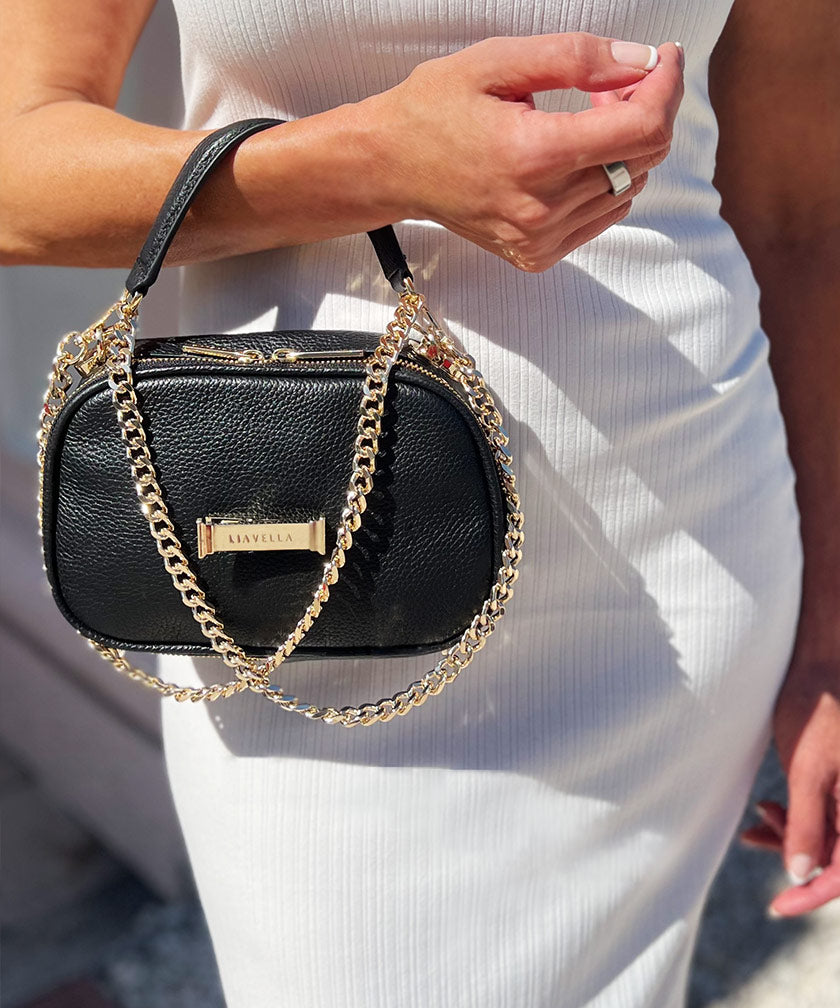 A person holding a noir AVAIA Petit, using the gold chain strap, twisted in a handheld style.