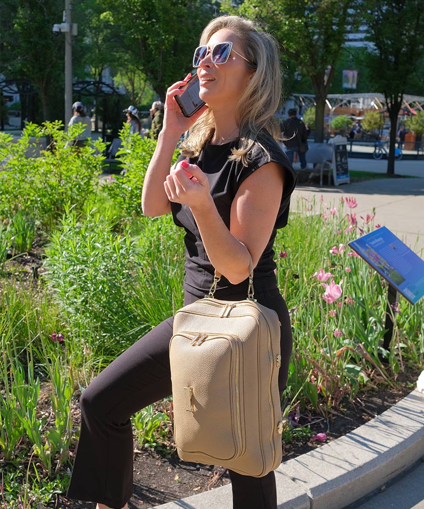 Woman talking on a phone with a crème caramel AVAIA bag, using a handheld strap, in an outdoor setting.