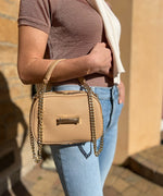 Load image into Gallery viewer, Person holding an crème caramel AVAIA Petit handbag with gold chain strap against a blurred background