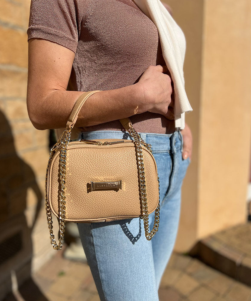 Person holding an crème caramel AVAIA Petit handbag with gold chain strap against a blurred background