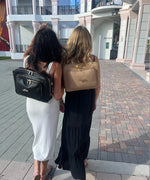 Load image into Gallery viewer, Two women walking together on a pavement, each wearing an AVAIA bag as backpacks, one crème caramel and noir.