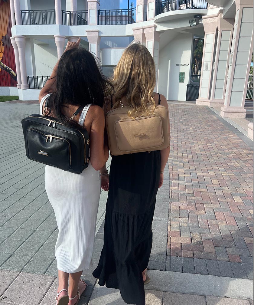 Two women walking together on a pavement, each wearing an AVAIA bag as backpacks, one crème caramel and noir.