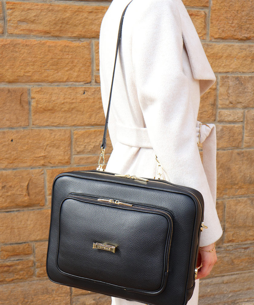 A person in a white coat, wearing the AVAIA bag over the shoulder against a brickwall backdrop.