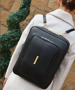Load image into Gallery viewer, A woman in a white coat with the AVAIA bag in Noir worn as a backpack, against a brickwall.
