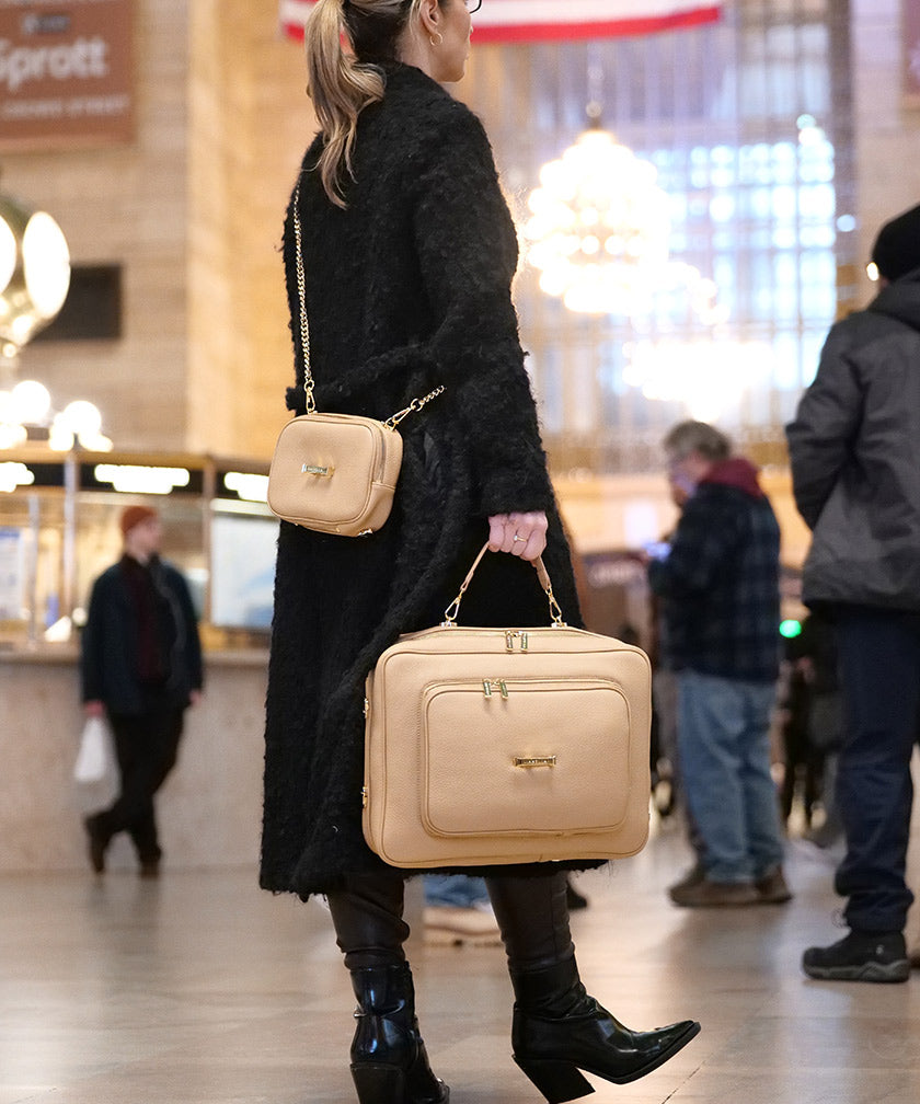 Woman holding a AVAIA crème caramel and wearing the AVAIA Petit in crème caramel crossbody in a busy indoor setting