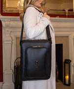Load image into Gallery viewer, Woman in a white coat wearing the AVAIA bag in Noir over the shoulder, in an elegant interior setting.
