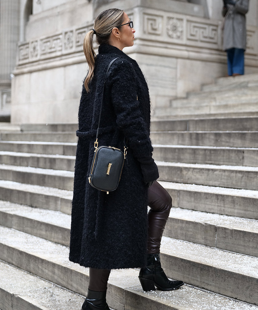 Woman in a black coat standing and a AVAIA Petit Noir with the fabric strap on stone steps with a building in the background