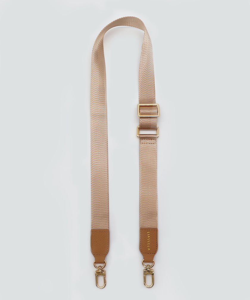 A wide fabric strap in creme caramel, with leather accents and gold hardware, against a white background.
