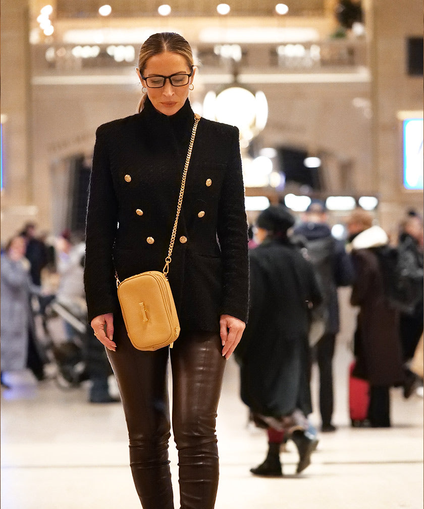 Woman in a black coat with gold buttons wearing the AVAIA Petit in crème caramel across her body, in a busy indoor setting