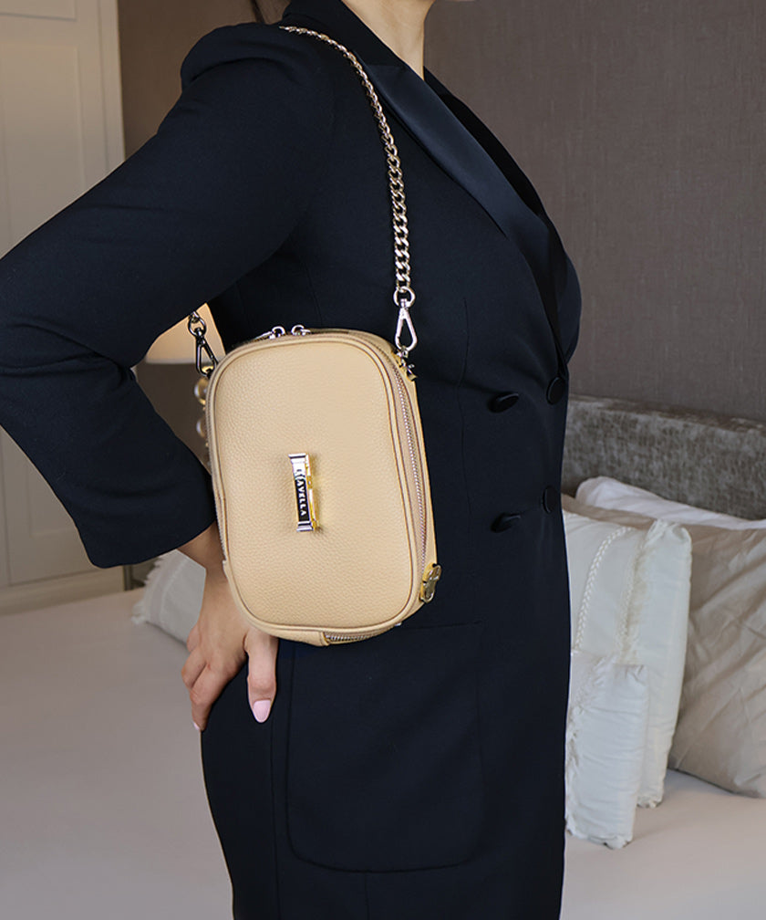 Person in a room wearing the avaia petit bag in creme caramel over the shoulder in vertical orientation. Showcasing the gold chain strap.