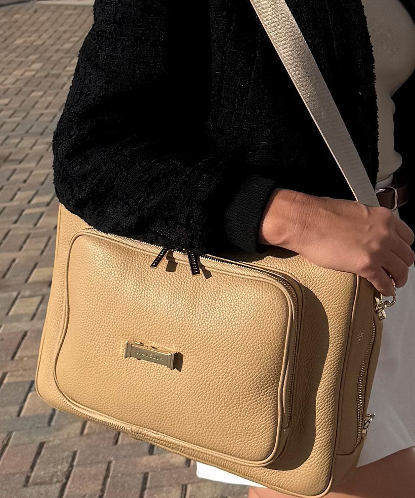 AVAIA creme caramel handbag held by a person with a black textured sleeve on a brick pavement background