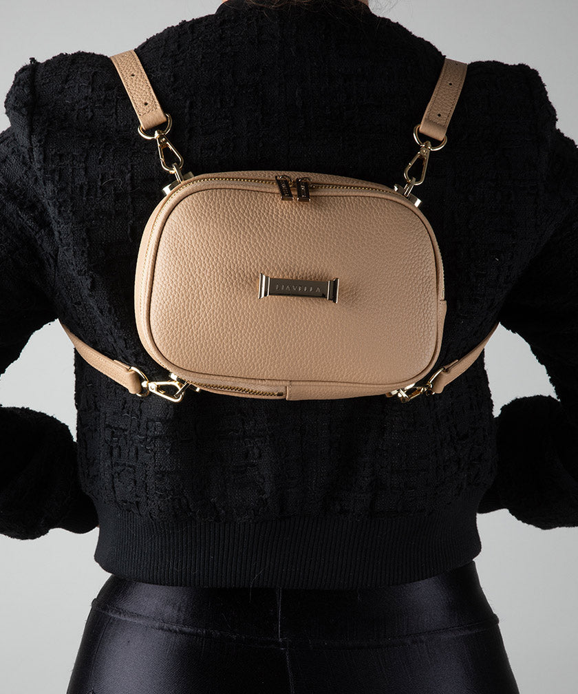 Person wearing a AVAIA Petit in crème caramel with two leather straps as a backpack with a black sweater and pants on a light gray background